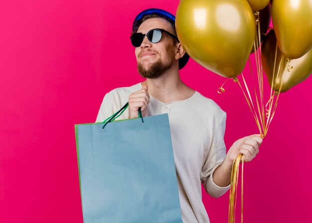 Pleased young handsome slavic party guy wearing party hat and sunglasses holding balloons and paper bags looking at side isolated on crimson background with copy space