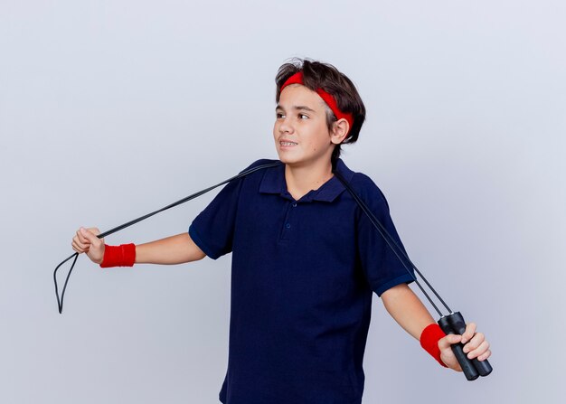 Pleased young handsome sporty boy wearing headband and wristbands with dental braces and jump rope around neck grabbing jump rope looking at side isolated on white background