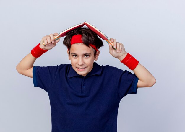 Pleased young handsome sporty boy wearing headband and wristbands with dental braces  touching head with ping pong rackets isolated on white wall