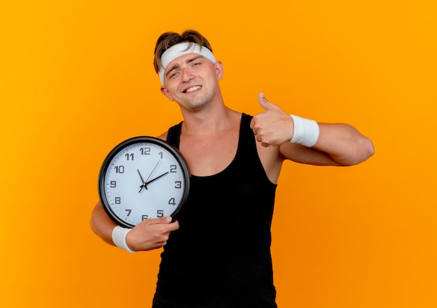 Pleased young handsome sporty man wearing headband and wristbands holding clock and showing thumb up isolated on orange wall