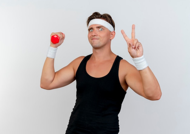 Pleased young handsome sporty man wearing headband and wristbands holding dumbbell and doing peace sign isolated on white wall