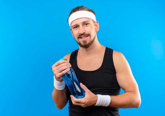 Pleased young handsome sporty man wearing headband and wristbands holding water bottle looking  isolated on blue space 