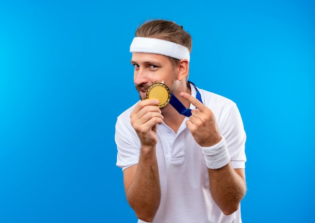 Pleased young handsome sporty man wearing headband and wristbands and medal around neck holding and pointing at medal isolated on blue space 