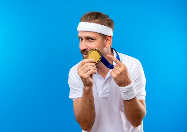 Довольный молодой красивый спортивный мужчина с повязкой на голову и браслетами и медалью на шее, держащей и указывая на медаль, изолированную на синем пространстве