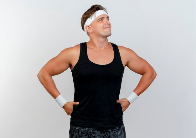 Pleased young handsome sporty man wearing headband and wristbands putting hands on waist and looking at side isolated on white wall