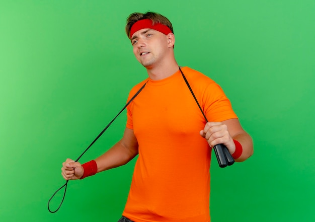 Pleased young handsome sporty man wearing headband and wristbands with jump rope around neck holding jump rope looking straight isolated on green wall
