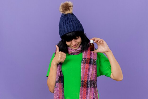 Довольная молодая больная кавказская девушка в зимней шапке и шарфе держит и смотрит на лекарство в стекле, показывая большой палец вверх, изолированный на фиолетовом фоне с копией пространства