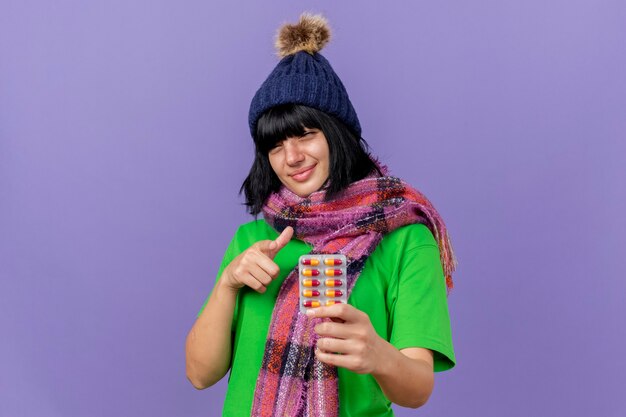 Довольная молодая больная кавказская девушка в зимней шапке и шарфе держит пачку капсул, смотрит и указывает на камеру, изолированную на фиолетовом фоне с копией пространства