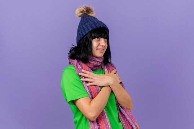 Pleased young ill caucasian girl wearing winter hat and scarf looking up keeping hands crossed on chest isolated on purple wall with copy space