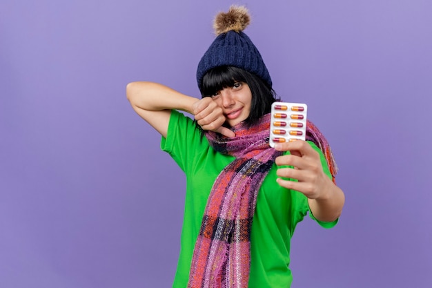 Free photo pleased young ill caucasian girl wearing winter hat and scarf stretching out pack of capsules showing thumb up looking at camera isolated on purple background with copy space