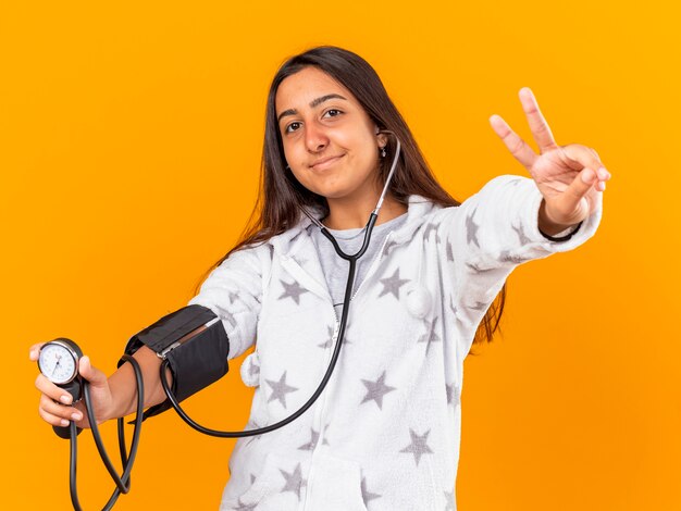 Pleased young ill girl measuring her own pressure with sphygmomanometer showing peace gesture isolated on yellow background