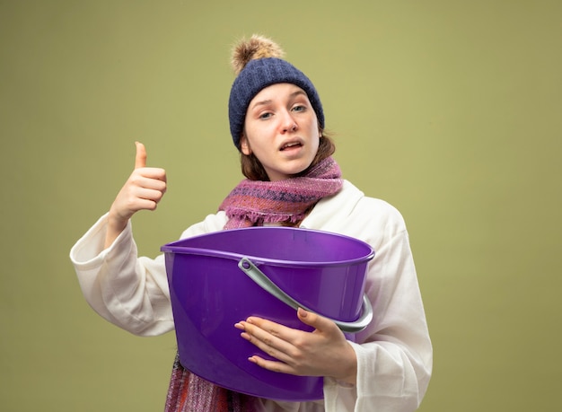 Felice giovane ragazza malata che indossa una veste bianca e cappello invernale con sciarpa che tiene secchio di plastica che mostra il pollice in alto isolato su verde oliva