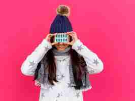 Free photo pleased young ill girl wearing winter hat with scarf covered eyes with pills isolated on pink