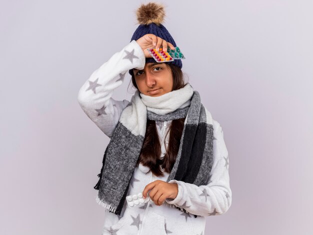 Pleased young ill girl wearing winter hat with scarf holding pills putting hand on forehead isolated on white background