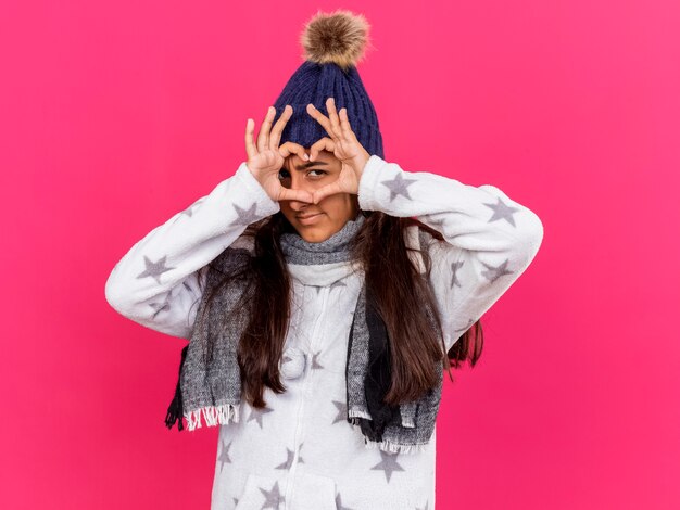 Free photo pleased young ill girl wearing winter hat with scarf showing heart gesture isolated on pink background