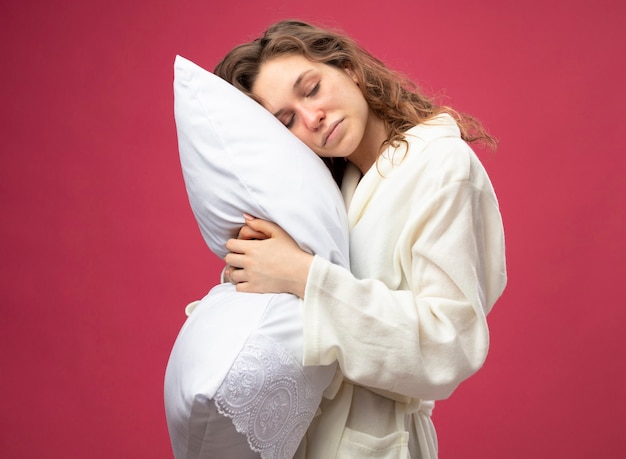 Pleased young ill girl with closed eyes wearing white robe hugged pillow isolated on pink