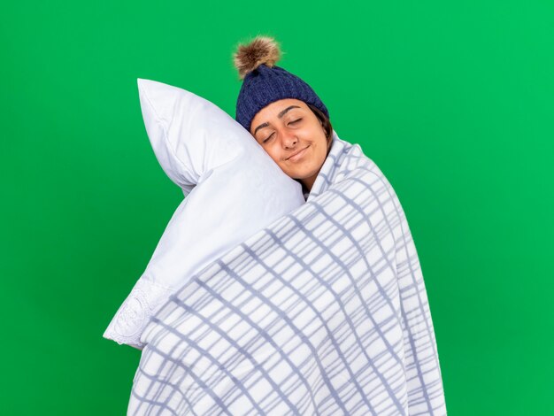 Pleased young ill girl with closed eyes wearing winter hat with scarf wrapped in plaid hugging pillow isolated on green background