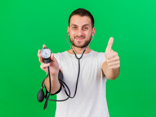 Pleased young ill man holding and wearing stethoscope showing thumb up isolated on green background