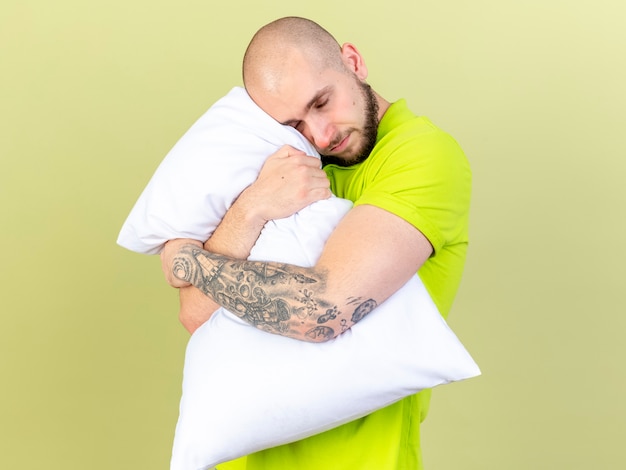 Pleased young ill man hugs pillow isolated on olive green wall