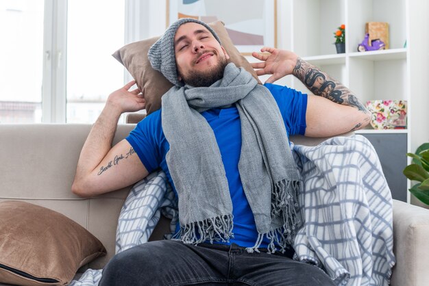 pleased young ill man wearing scarf and winter hat sitting on sofa in living room holding pillow behind his head resting head on it with closed eyes