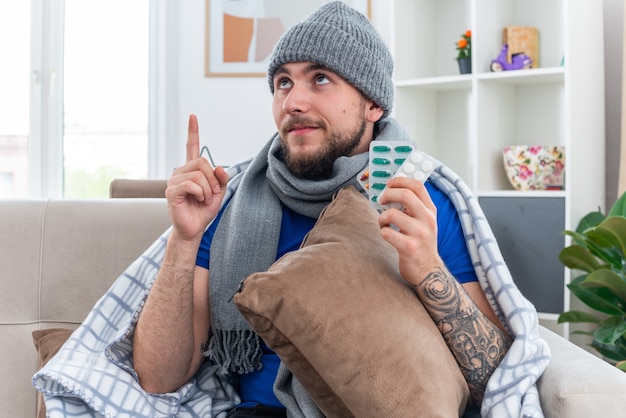 毛布に包まれたスカーフと冬の帽子を身に着けている若い病気の人を喜ばせるリビングルームのソファに座って枕を持って丸薬のパックを見て上向きに