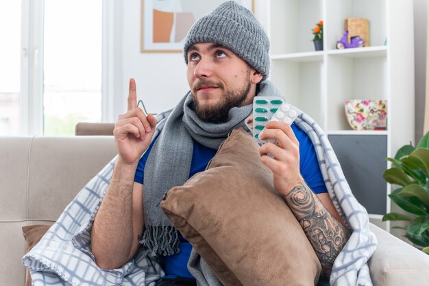 Довольный молодой больной человек в шарфе и зимней шапке, завернутый в одеяло, сидит на диване в гостиной, держа подушку с пачками таблеток, смотрит и указывает вверх