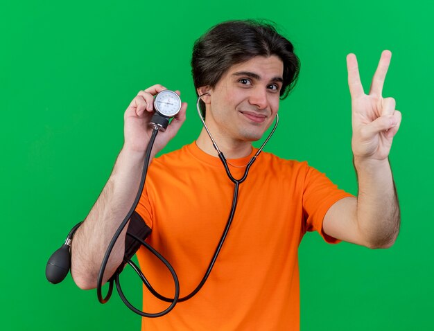 Pleased young ill man wearing stethoscope holding sphygmomanometer showing peace gesture isolated on green