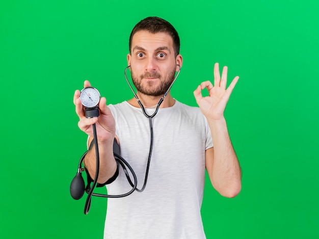 聴診器を身に着けて、緑の背景に分離された血圧計で自分の圧力を測定する若い病気の人を喜ばせる