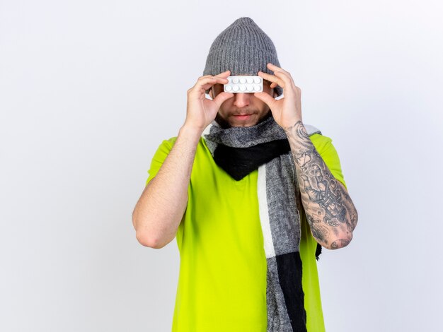 冬の帽子とスカーフを身に着けている幸せな若い病気の人は、白い壁に隔離された目の前に医療錠剤のパックを保持します