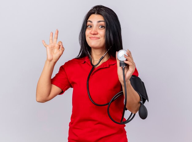 Pleased young ill woman wearing stethoscope looking at front measuring her pressure with sphygmomanometer doing ok sign isolated on white wall