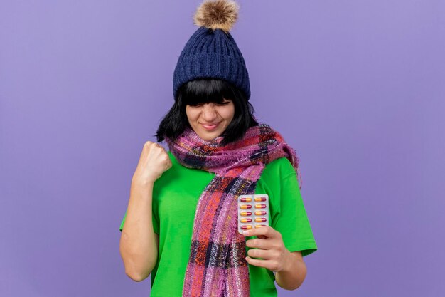 Pleased young ill woman wearing winter hat and scarf holding pack of capsules doing yes gesture isolated on purple wall with copy space