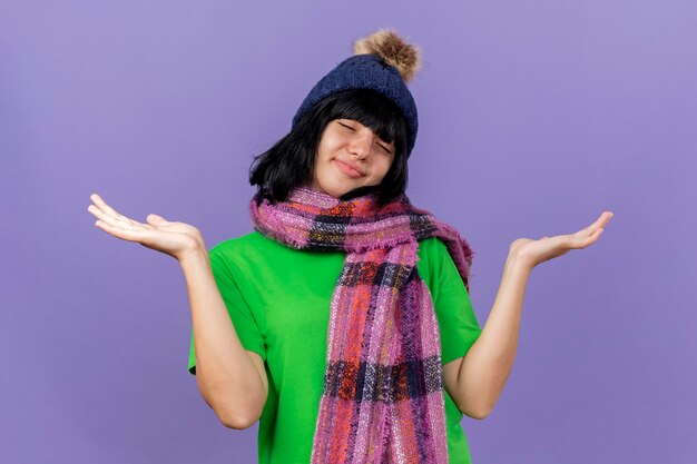 Pleased young ill woman wearing winter hat and scarf showing empty hands with closed eyes isolated on purple wall