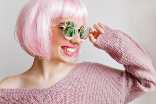 Pleased young lady in wig smiling during indoor photoshoot. Close-up portrait of laughing stylish woman with pink hair posing in sparkle glasses.