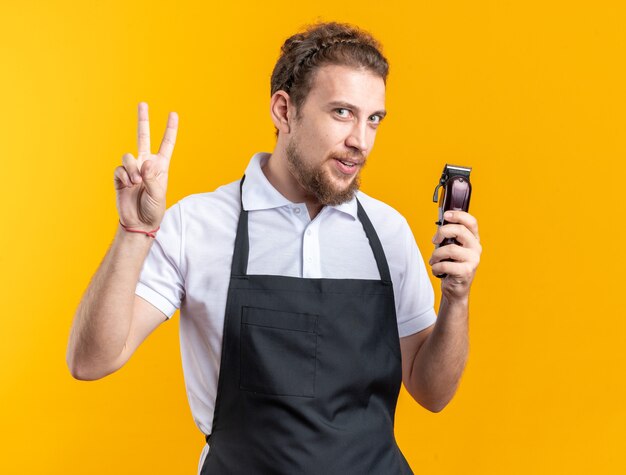 Pleased young male barber wearing uniform holding hair clippers showing peace gesture isolated on yellow wall