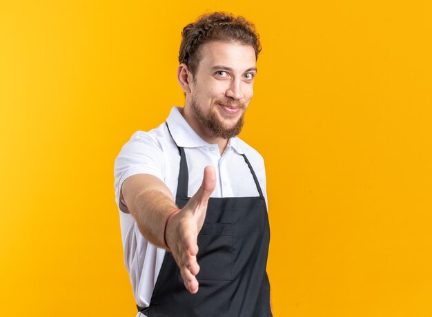 Pleased young male barber wearing uniform holding out hand at camera isolated on yellow wall