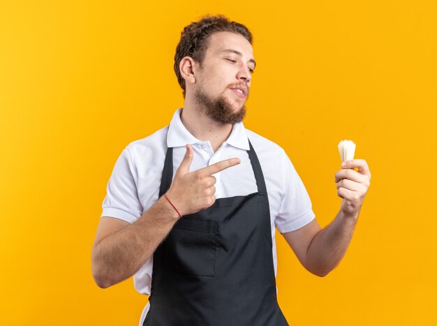 Pleased young male barber wearing uniform holding and points at shaving brush isolated on yellow wall