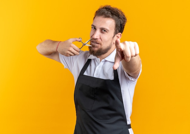 Pleased young male barber wearing uniform holding scissors points at camera isolated on yellow wall