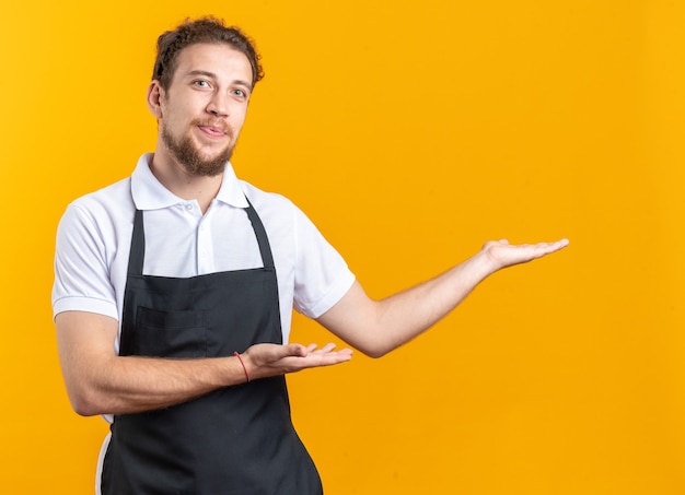 Pleased young male barber wearing uniform points at side isolated on yellow wall with copy space