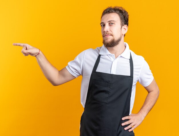 Pleased young male barber wearing uniform points at side putting hand on hip isolated on yellow wall