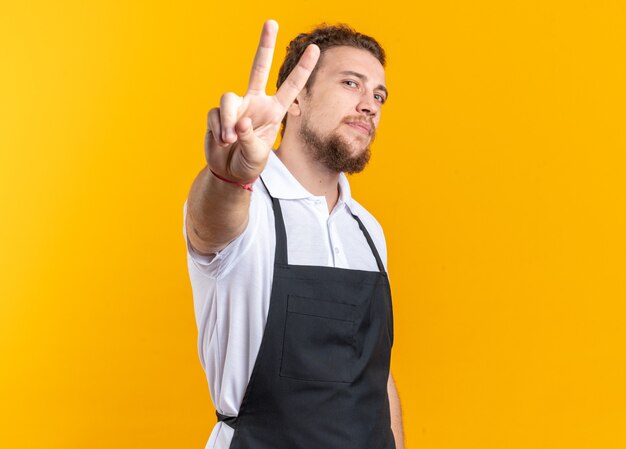 Pleased young male barber wearing uniform showing peace gesture isolated on yellow wall