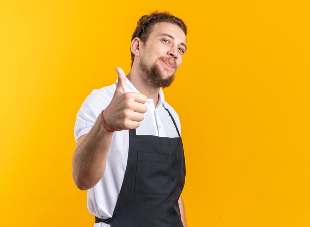 Pleased young male barber wearing uniform showing thumb up isolated on yellow wall