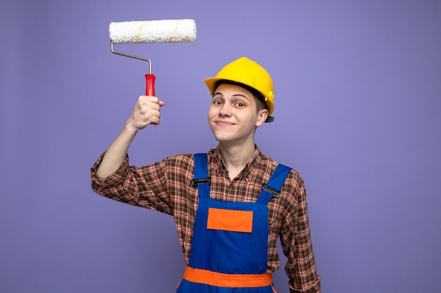 Pleased young male builder wearing uniform holding roller brush 