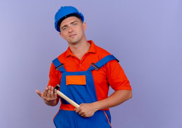 Free photo pleased young male builder wearing uniform and safety helmet holding hammer