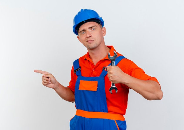 Free photo pleased young male builder wearing uniform and safety helmet holding wrench and points at side on white
