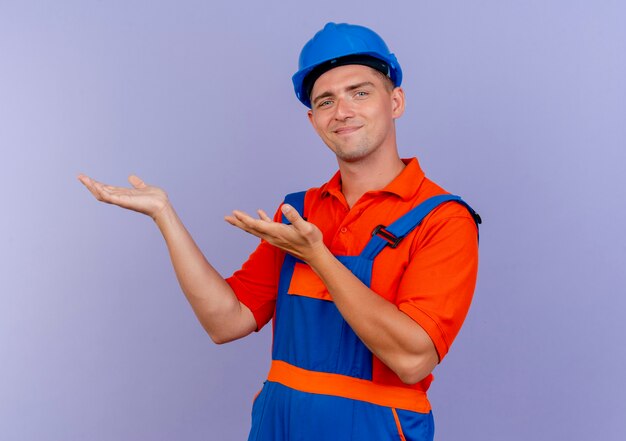 Pleased young male builder wearing uniform and safety helmet pretending holding something on purple