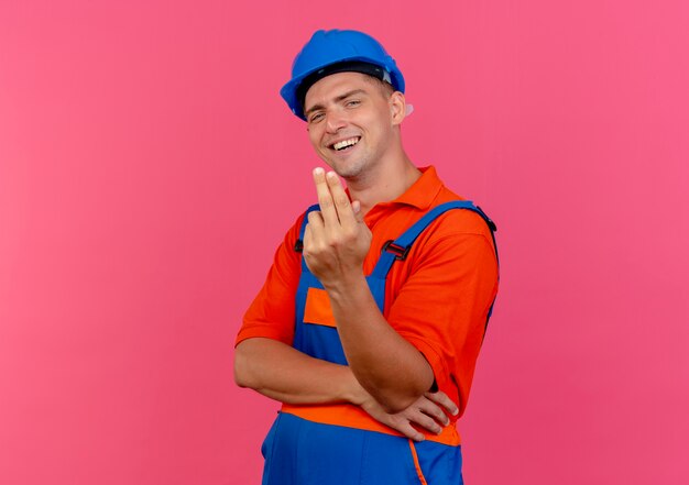 Pleased young male builder wearing uniform and safety helmet showing two on pink