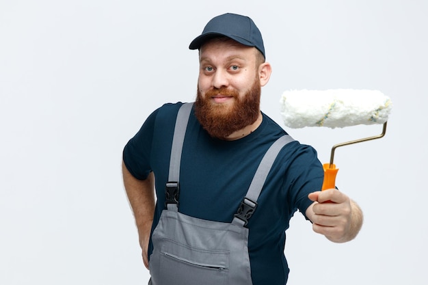 Pleased young male construction worker wearing cap and uniform stretching paint roller out towards camera keeping hand on waist looking at camera isolated on white background