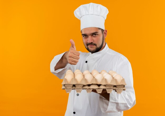 卵のカートンを保持し、オレンジ色のスペースで隔離された親指を見せてシェフの制服を着た若い男性料理人を喜ばせる