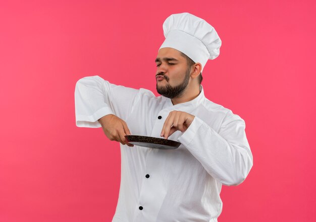 Pleased young male cook in chef uniform holding frying pan and putting fingers on it with closed eyes isolated on pink space