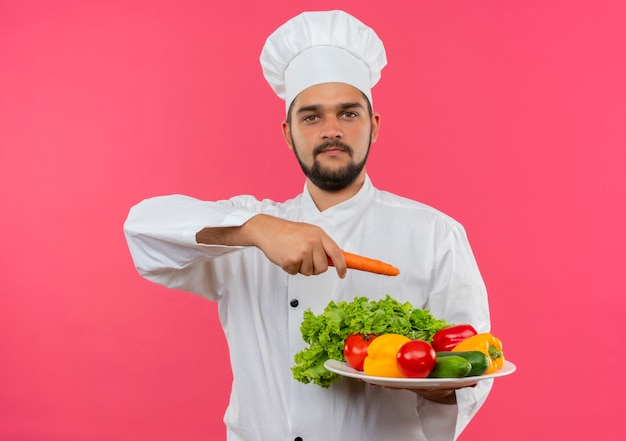 Pleased young male cook in chef uniform holding plate of vegetables and pointing with carrot at it isolated on pink space